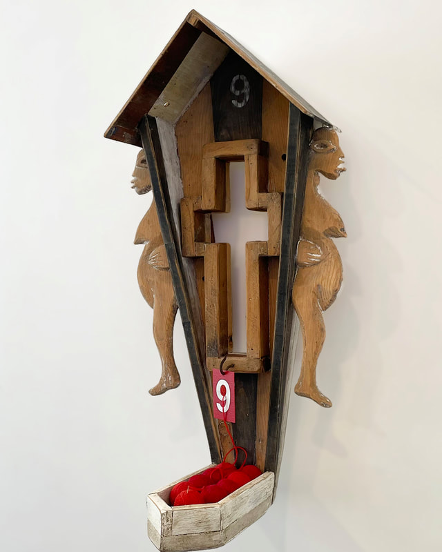 3D artwork, with hand carved pregnant female fiigures flanking a house shape with a cross cut in it and balls of red thread.