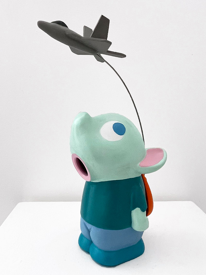 cartoonish sculpture of a child-like creature with a snout/nose and pink animal-like ears with a toy fighter jet arcing overhead 