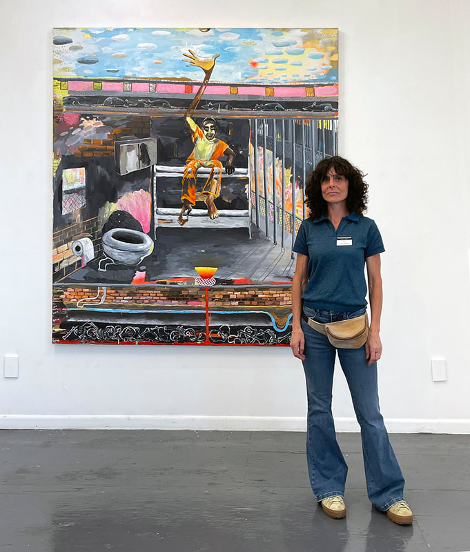 woman with shoulder length curly hair in jeans and a tshirt standing in fron of a painting of a man sitting ina prison cell with one hand reaching up and out through the roof to the sky
