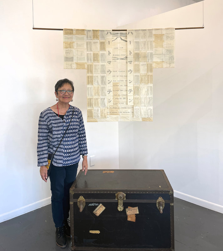 Woman with short dark hair and glasses standing next to a trunk with an artwork shaped like a happi hanging above it.
