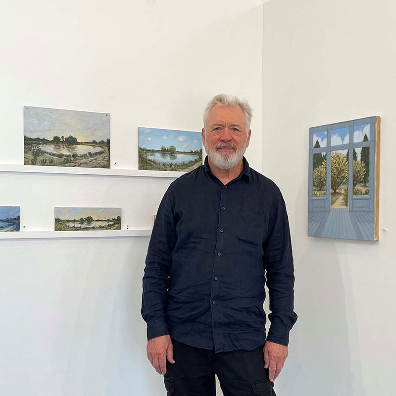 Man in dark button up shirt standing in front of a series of small paintings on cardboard of a pond with trees in the distance
