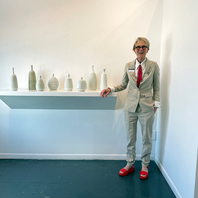 woman with short hair in a beige linen suit with red tie and red sandals standing near a shelf with hand made ceramic bottles