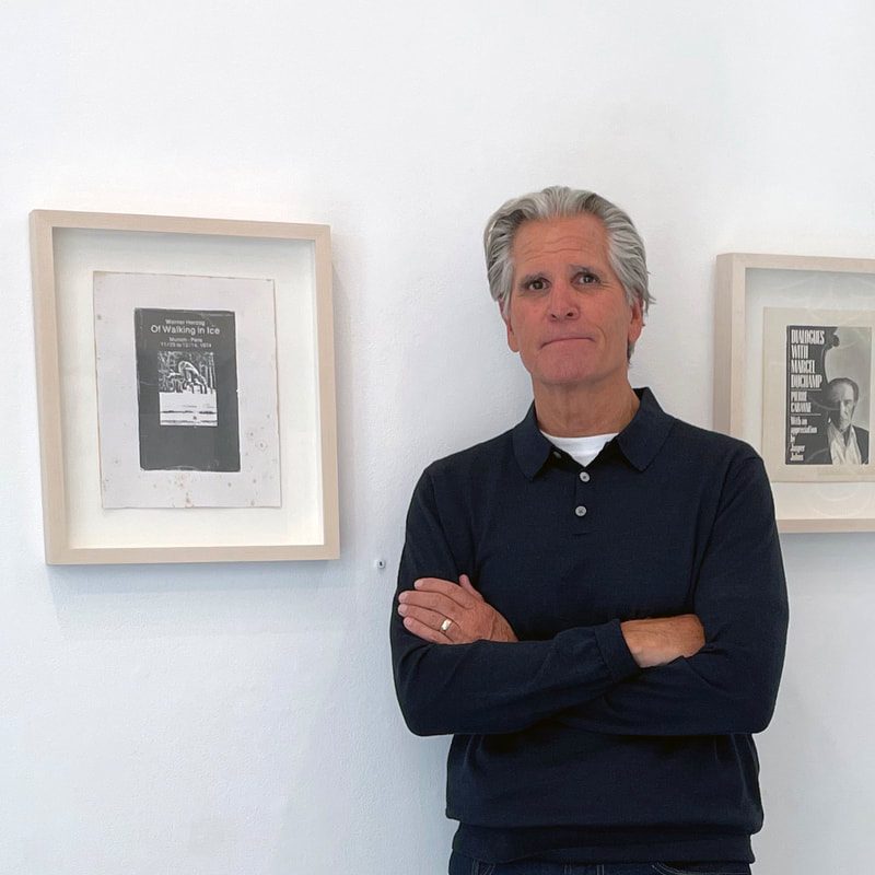 Man with gray hair in a long sleeved shirt, arms folded staning next to two framed drawings of book covers, one of a book abbout Werner herzog and the other a book about Duchamp.