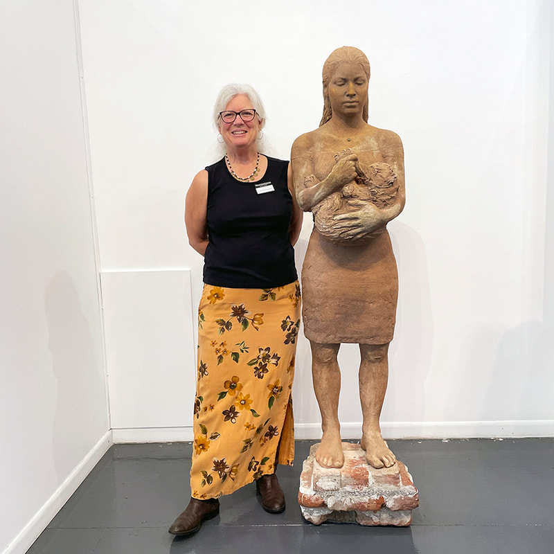 woman with whte hair in a black top and yellow long skirt standing next to a sculputre of a life size woman holding a bear cub