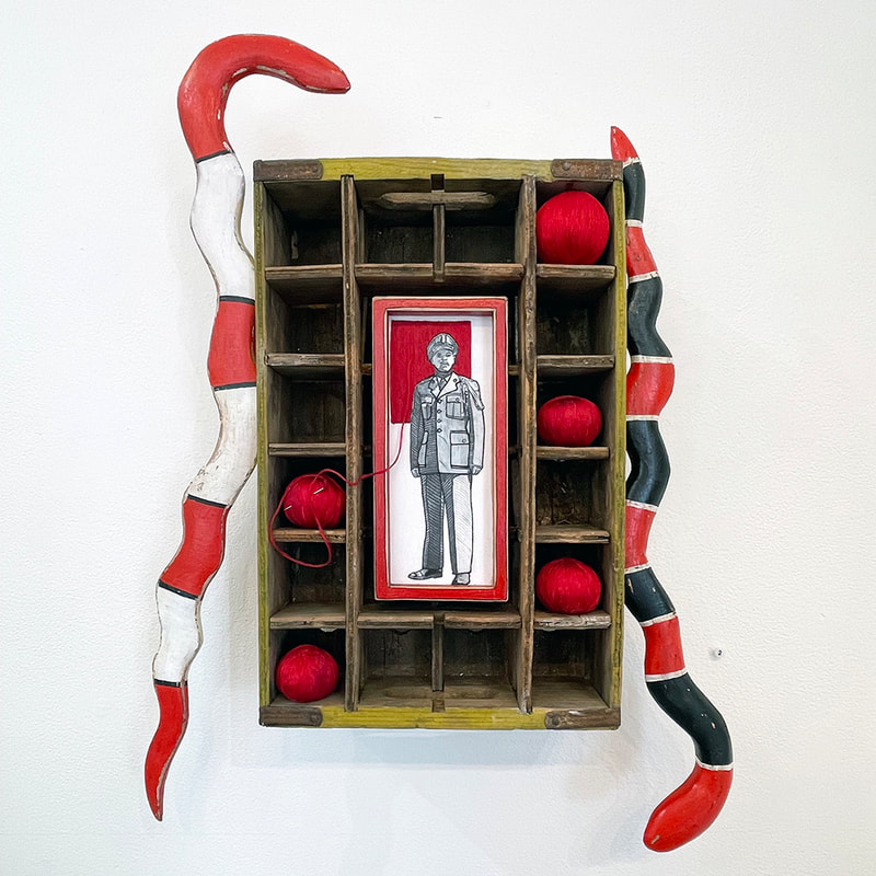 Artwork with graphite portrait of a black US Military soldier attached to a wooden crate with balls of red thread and carved wood snakes