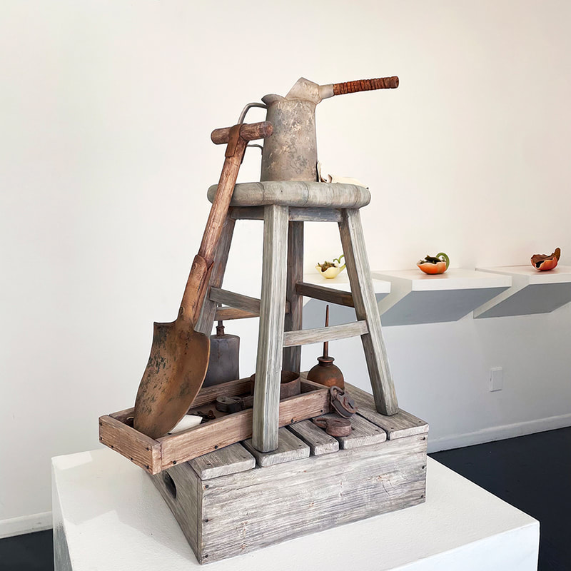 porcelain sculpture that looks like a watering can on top of a wooden stool on top of a wooden crate with a shovel leaning against the stool