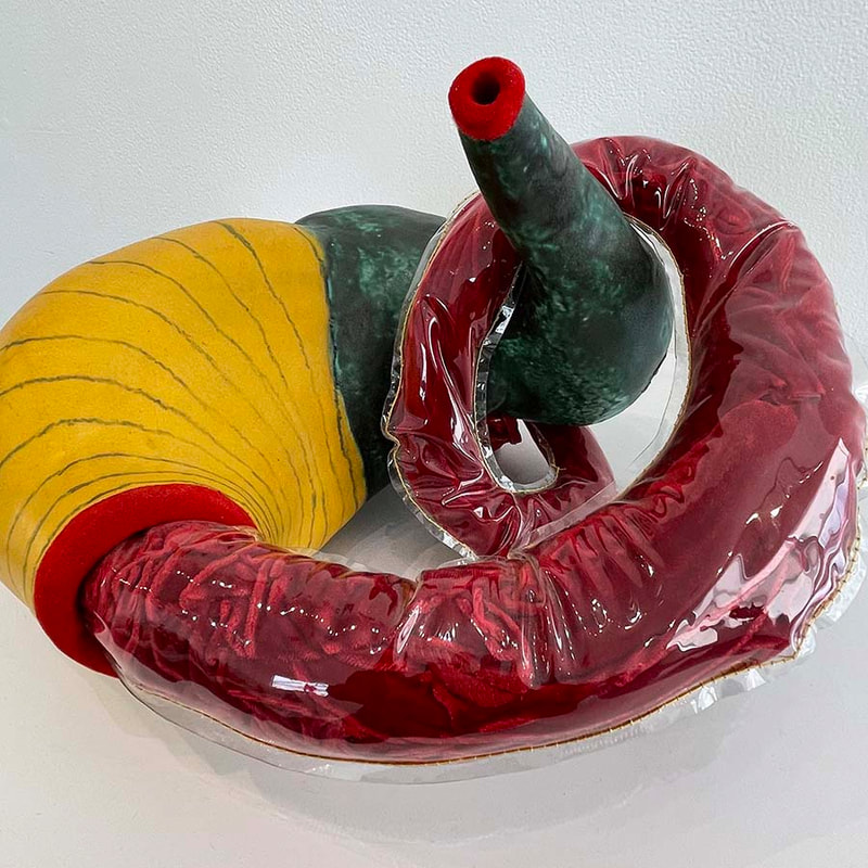 Curvy sculpture made of ceramic amorphic shapes with a clear plastic sleeve encasing a red velvet child's dress looping around one end of it