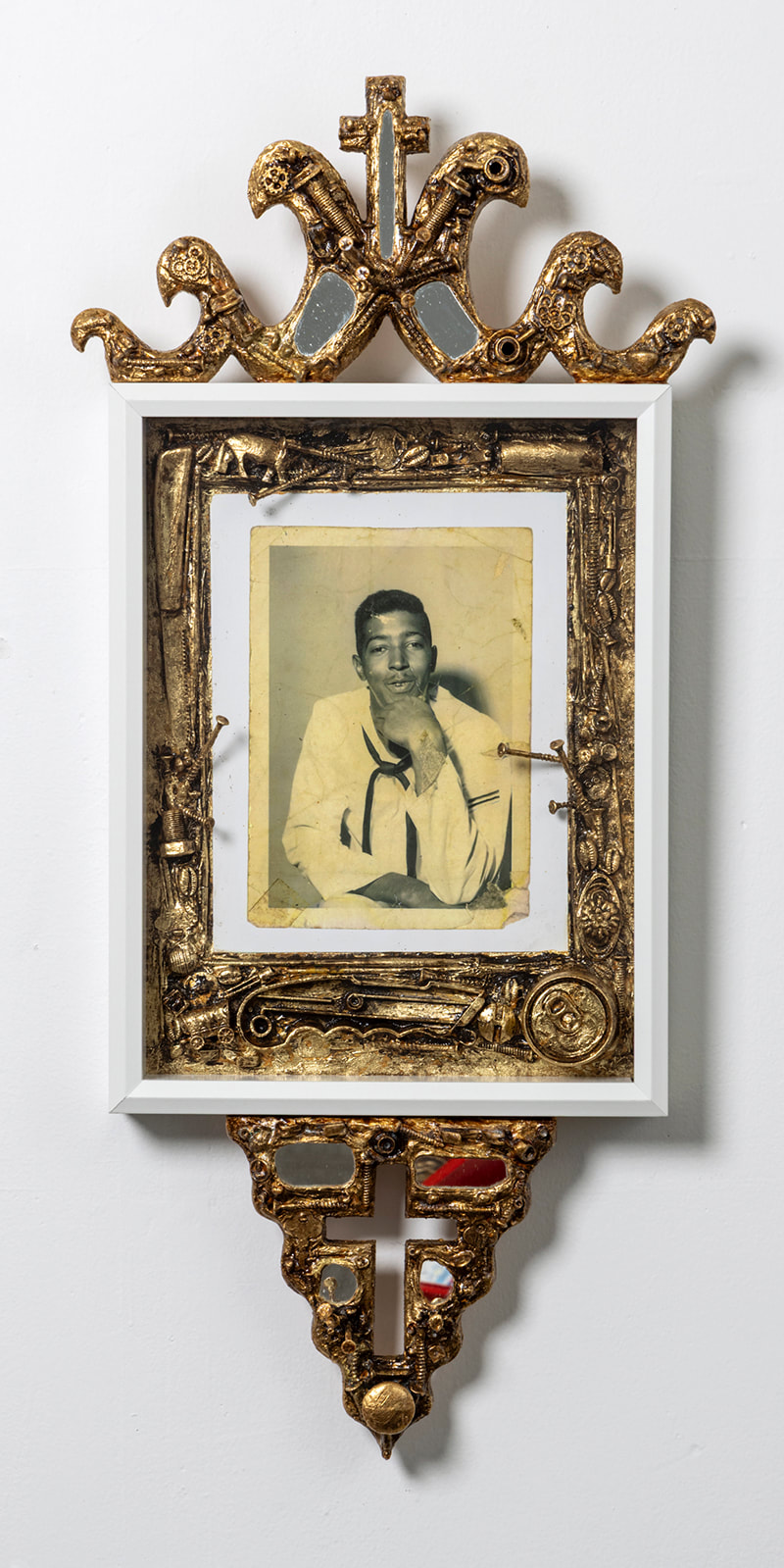 photo portrait of a Black sailor in a frame bordered by gold leafed screws, nails and d hardware and with a gold leafed cross and hand carved flames on top