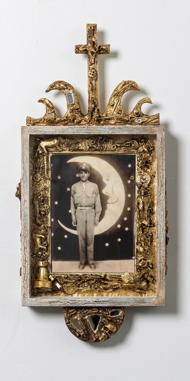 photo portrait of a Black soldier standing in fron of a large paper slivered moon in a frame bordered by gold leafed trinkets and hardware and with a cross on topPicture